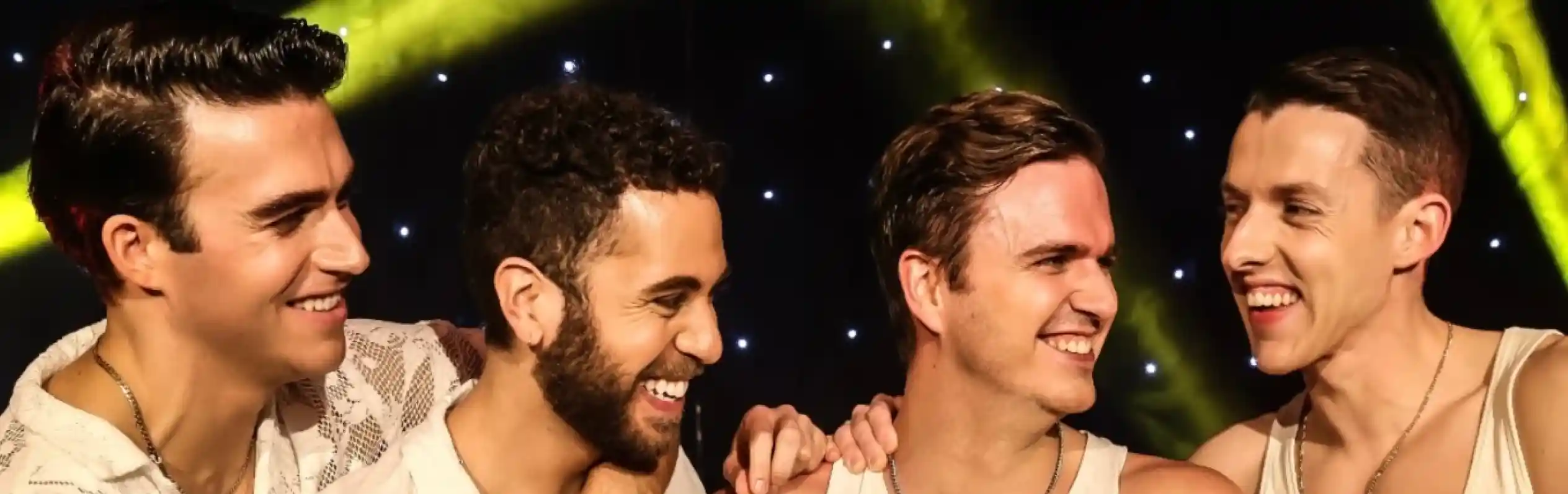 Four men dressed in casual white outfits laugh and interact with each other on stage, illuminated by colorful lights. They display camaraderie and joy as they smile at one another.