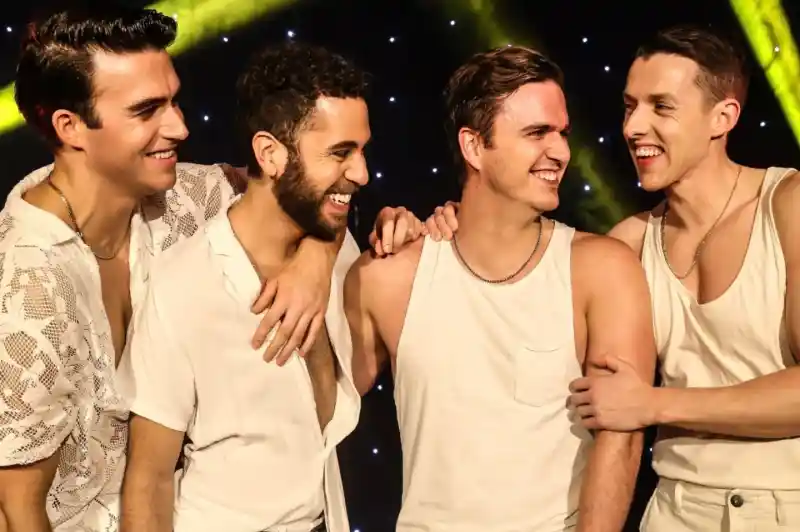 Four men dressed in casual white outfits laugh and interact with each other on stage, illuminated by colorful lights. They display camaraderie and joy as they smile at one another.