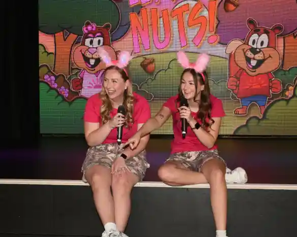 Two women in matching pink shirts and camo skirts sit on a stage. They each hold a microphone and wear fluffy pink ear headbands. In the background, colorful cartoon characters and the phrase "We're Nuts!" are displayed on a large screen. The women are laughing and interacting with each other.