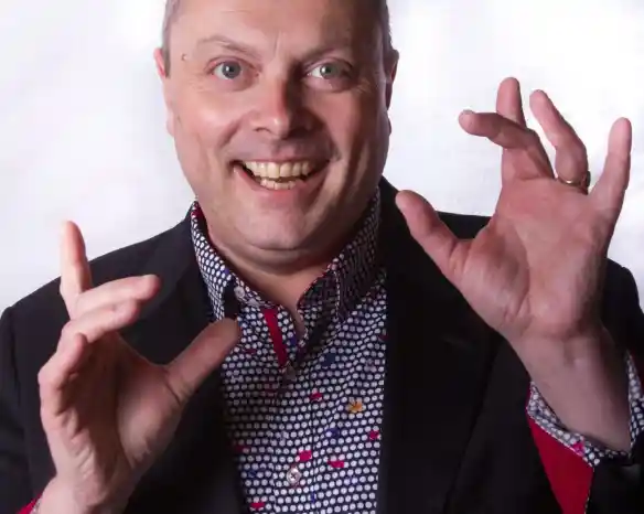 A middle-aged man with a cheerful expression poses with hands raised and gesturing. He wears a dark blazer over a colorful, patterned shirt.