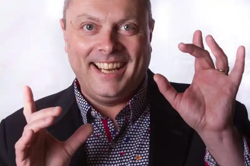 A middle-aged man with a cheerful expression poses with hands raised and gesturing. He wears a dark blazer over a colorful, patterned shirt.