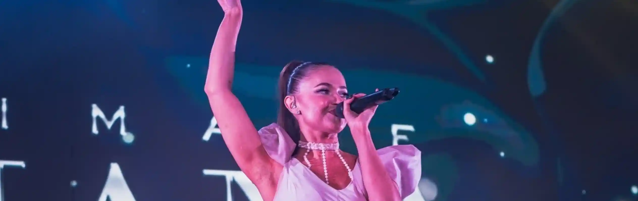 A female singer performs on stage, wearing a white outfit with a skirt. She has her right arm raised and is holding a microphone. A male musician with a bass guitar stands behind her, blending into an illuminated background.
