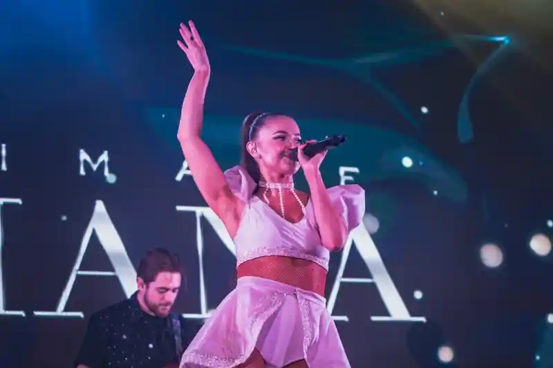 A female singer performs on stage, wearing a white outfit with a skirt. She has her right arm raised and is holding a microphone. A male musician with a bass guitar stands behind her, blending into an illuminated background.