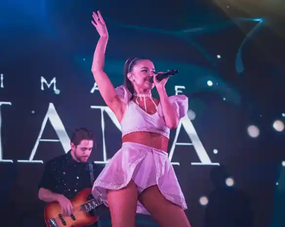 A female singer performs on stage, wearing a white outfit with a skirt. She has her right arm raised and is holding a microphone. A male musician with a bass guitar stands behind her, blending into an illuminated background.