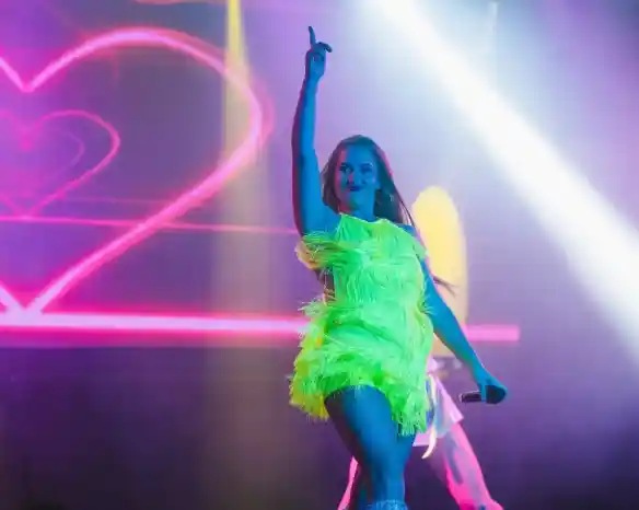 A performer in a vibrant neon green outfit with feathers strikes a dynamic pose on stage, raising one finger in the air. Colorful lights and a heart-shaped backdrop enhance the energetic atmosphere.