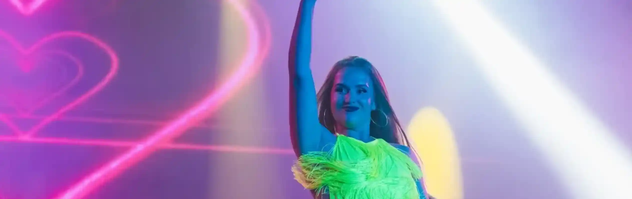 A performer in a vibrant neon green outfit with feathers strikes a dynamic pose on stage, raising one finger in the air. Colorful lights and a heart-shaped backdrop enhance the energetic atmosphere.