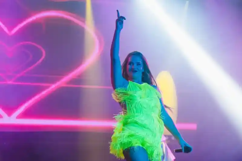 A performer in a vibrant neon green outfit with feathers strikes a dynamic pose on stage, raising one finger in the air. Colorful lights and a heart-shaped backdrop enhance the energetic atmosphere.