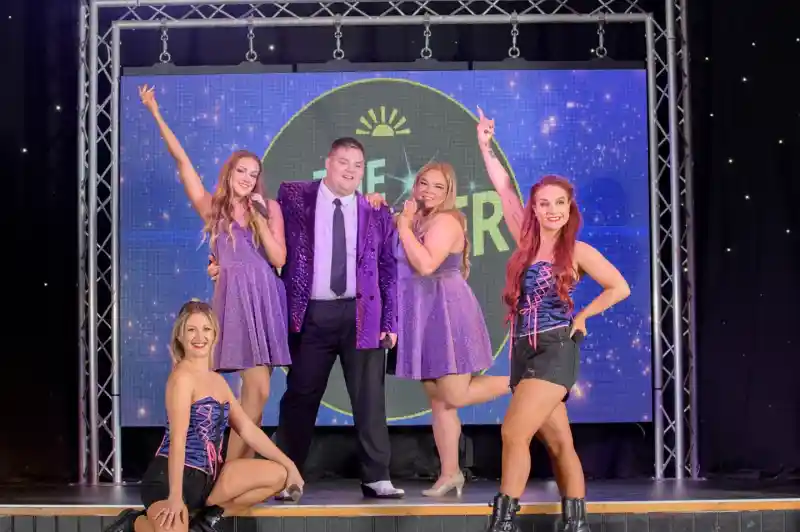 A group of five performers posing on a stage. Two women in matching sparkly purple dresses stand with a man in a shiny purple jacket and black trousers in front of a lit-up backdrop. Two other women in black shorts, fishnet tights, and colorful corsets are kneeling and standing at the front corners of the stage. The background features a large circular logo with stars and the text 'THE STAR'.