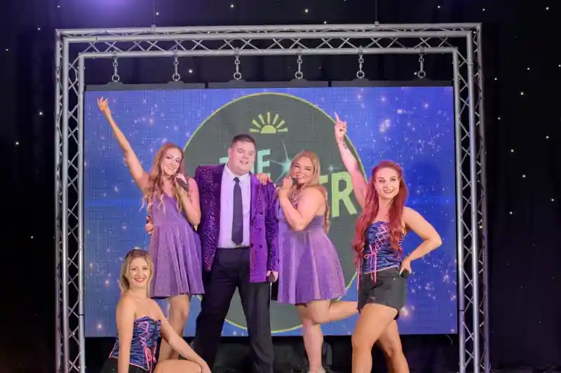 A group of five performers posing on a stage. Two women in matching sparkly purple dresses stand with a man in a shiny purple jacket and black trousers in front of a lit-up backdrop. Two other women in black shorts, fishnet tights, and colorful corsets are kneeling and standing at the front corners of the stage. The background features a large circular logo with stars and the text 'THE STAR'.