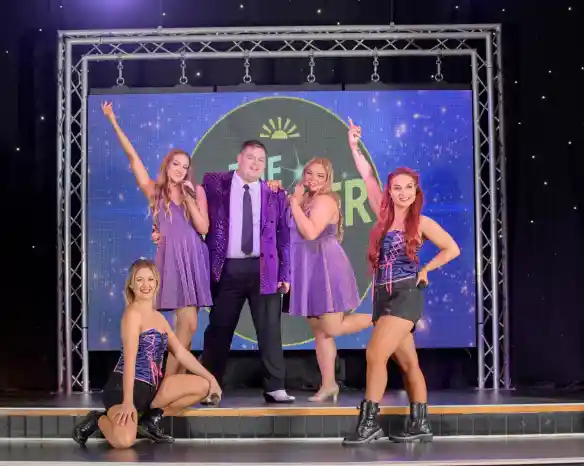 A group of five performers posing on a stage. Two women in matching sparkly purple dresses stand with a man in a shiny purple jacket and black trousers in front of a lit-up backdrop. Two other women in black shorts, fishnet tights, and colorful corsets are kneeling and standing at the front corners of the stage. The background features a large circular logo with stars and the text 'THE STAR'.