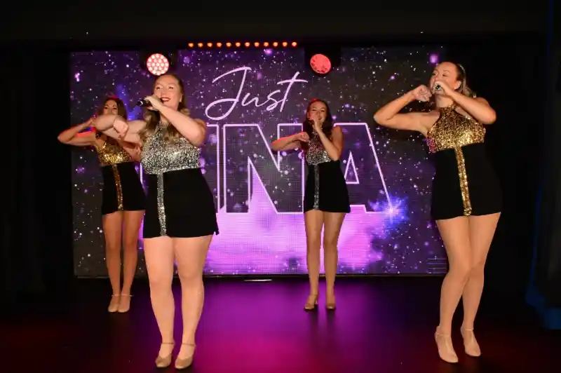 Four dancers perform on stage, wearing shiny, coordinated outfits with sequins, and each holds a microphone. The backdrop features the text "Justina" in a starry design, with colorful stage lights illuminating the scene.
