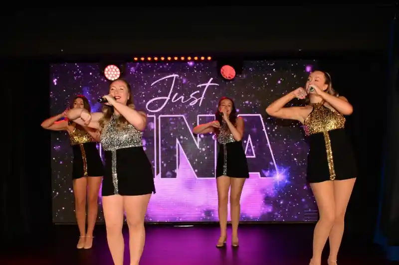 Four dancers perform on stage, wearing shiny, coordinated outfits with sequins, and each holds a microphone. The backdrop features the text "Justina" in a starry design, with colorful stage lights illuminating the scene.