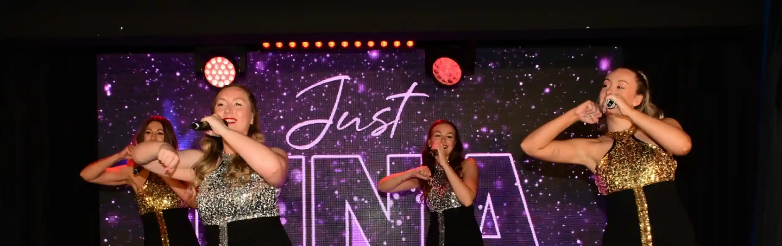 Four dancers perform on stage, wearing shiny, coordinated outfits with sequins, and each holds a microphone. The backdrop features the text "Justina" in a starry design, with colorful stage lights illuminating the scene.