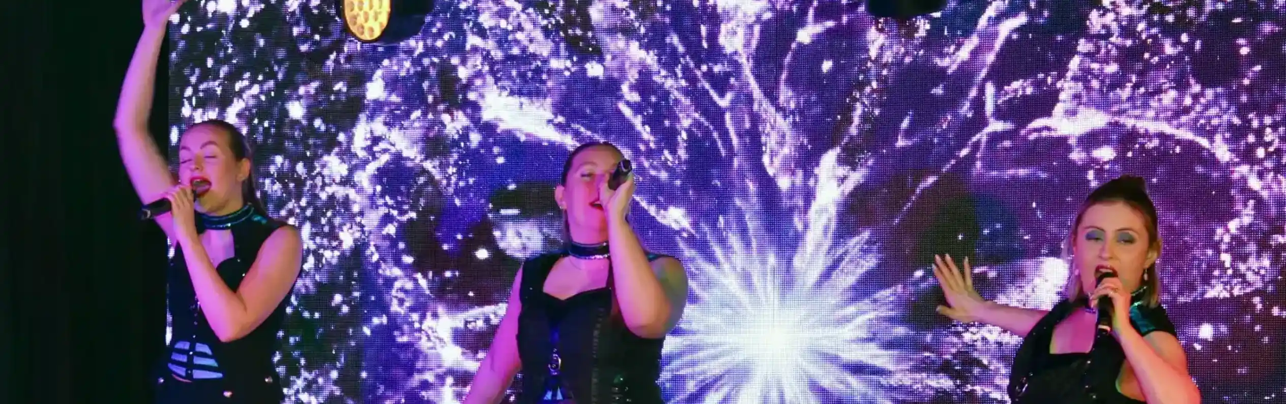 Three women perform on stage, dressed in black outfits with cut-out designs and combat boots. They are singing into microphones, with one raising her arm. Behind them is a vibrant, glowing background of swirling purple and white patterns.