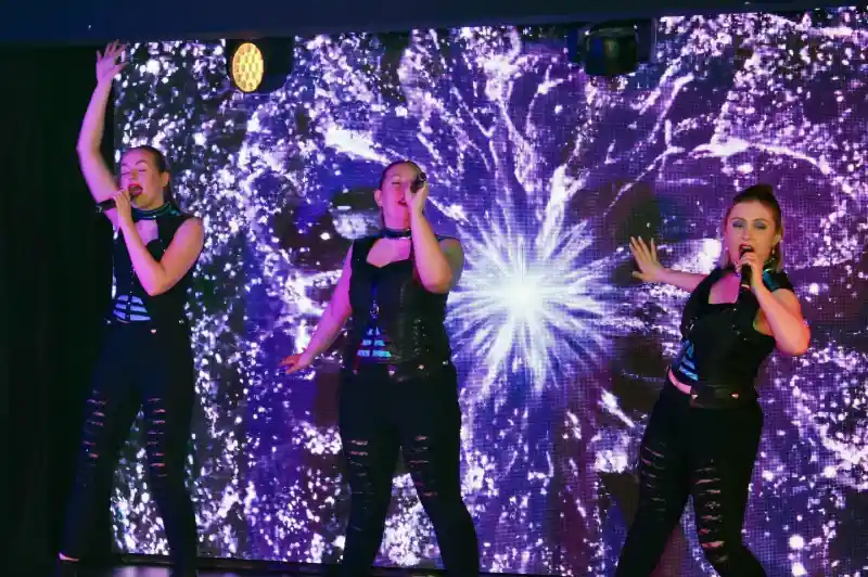 Three women perform on stage, dressed in black outfits with cut-out designs and combat boots. They are singing into microphones, with one raising her arm. Behind them is a vibrant, glowing background of swirling purple and white patterns.