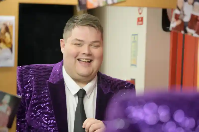 A smiling man in a sparkly purple blazer and black tie stands in a room with colorful posters on the wall. He appears to be adjusting his tie and is reflected in a mirror in the foreground.