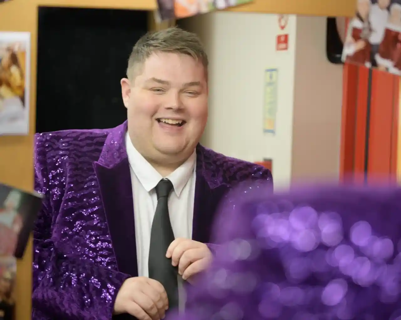 A smiling man in a sparkly purple blazer and black tie stands in a room with colorful posters on the wall. He appears to be adjusting his tie and is reflected in a mirror in the foreground.