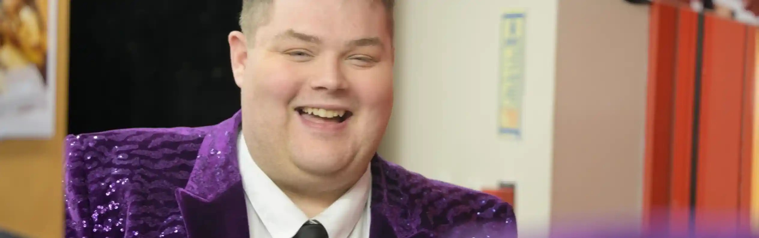 A smiling man in a sparkly purple blazer and black tie stands in a room with colorful posters on the wall. He appears to be adjusting his tie and is reflected in a mirror in the foreground.