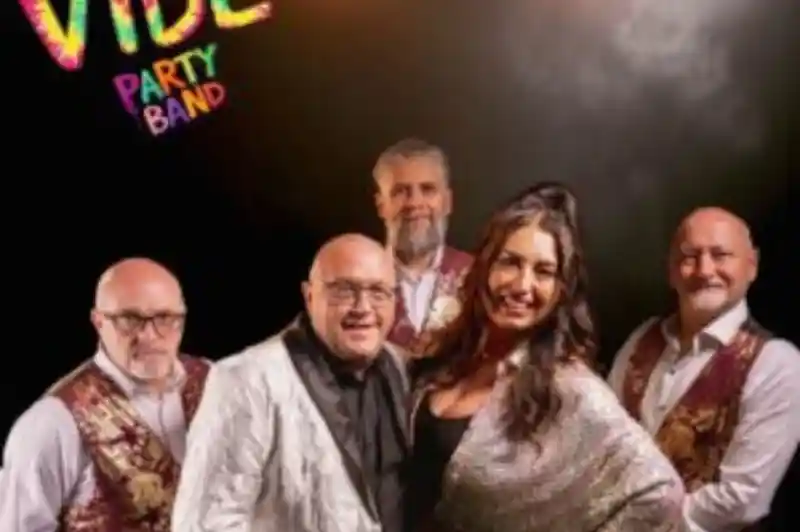 A party band poses together, featuring five members in stylish outfits. The front includes a woman in a sparkling top and a man in a sequined jacket, while the others wear vests. Colorful text in the corner reads "VIBE PARTY BAND." Bright stage lights create a festive atmosphere.