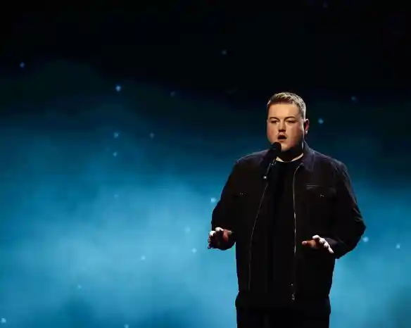 A man stands on stage with a microphone, wearing a black jacket and shirt. He appears to be performing, with a softly lit blue background featuring stars.