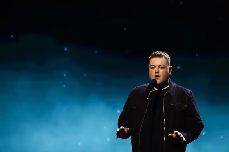 A man stands on stage with a microphone, wearing a black jacket and shirt. He appears to be performing, with a softly lit blue background featuring stars.