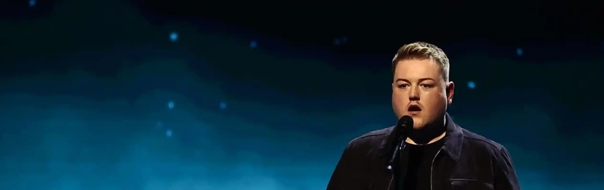 A man stands on stage with a microphone, wearing a black jacket and shirt. He appears to be performing, with a softly lit blue background featuring stars.