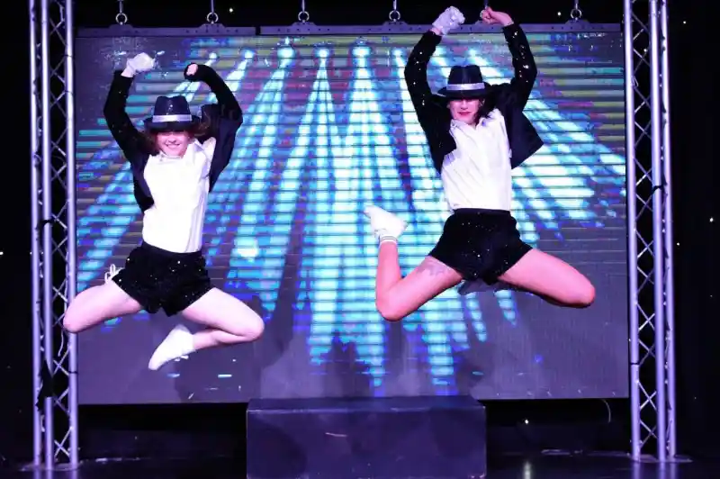 Two dancers in matching outfits perform a synchronized jump on stage, showcasing energetic choreography. The backdrop features dynamic colorful lights, enhancing the lively atmosphere of the performance.