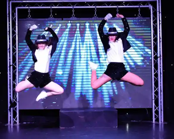 Two dancers in matching outfits perform a synchronized jump on stage, showcasing energetic choreography. The backdrop features dynamic colorful lights, enhancing the lively atmosphere of the performance.