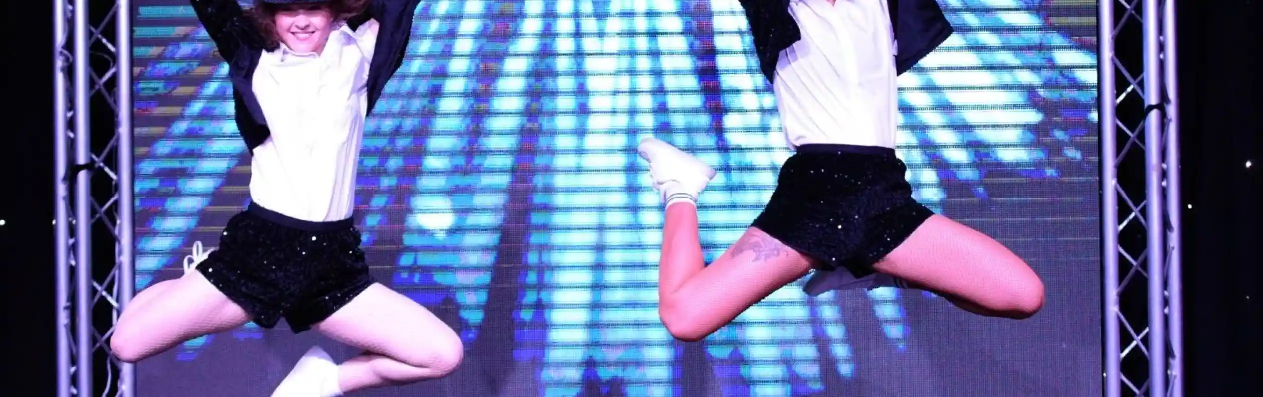 Two dancers in matching outfits perform a synchronized jump on stage, showcasing energetic choreography. The backdrop features dynamic colorful lights, enhancing the lively atmosphere of the performance.