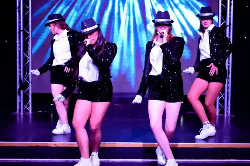Four performers in stylish black and white outfits with hats and sequins dance on stage. They are engaged in a synchronized routine, exuding energy and confidence, with colorful lights in the background.