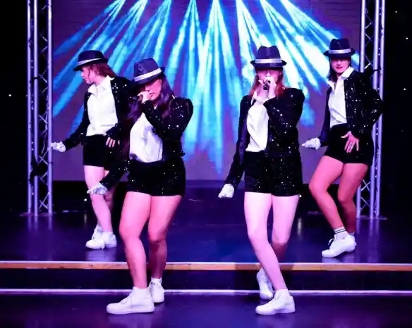 Four performers in stylish black and white outfits with hats and sequins dance on stage. They are engaged in a synchronized routine, exuding energy and confidence, with colorful lights in the background.