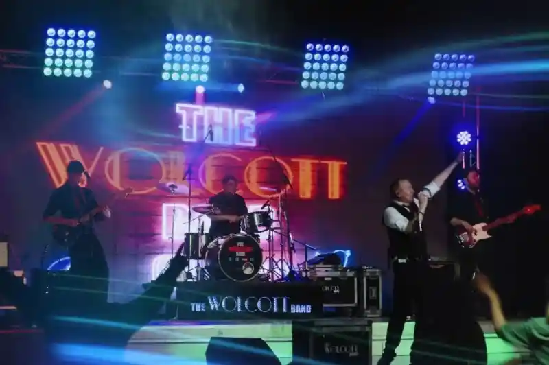 A live band performance with a lead singer on stage, energetically singing and gesturing. The band includes a drummer and two guitarists, all under colorful stage lights. The backdrop features the name "The Wolcott Band" in bright letters. Audience members are seen enjoying the performance.