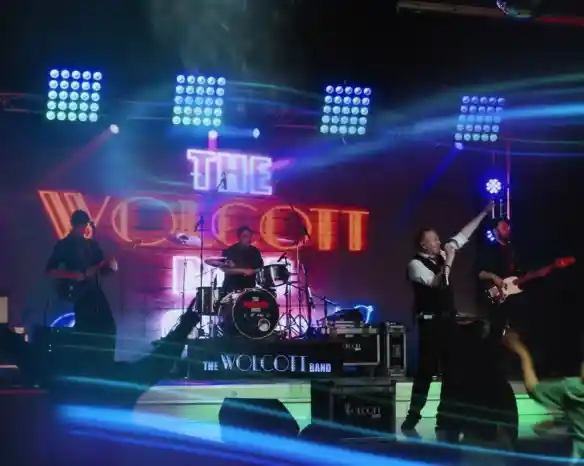 A live band performance with a lead singer on stage, energetically singing and gesturing. The band includes a drummer and two guitarists, all under colorful stage lights. The backdrop features the name "The Wolcott Band" in bright letters. Audience members are seen enjoying the performance.