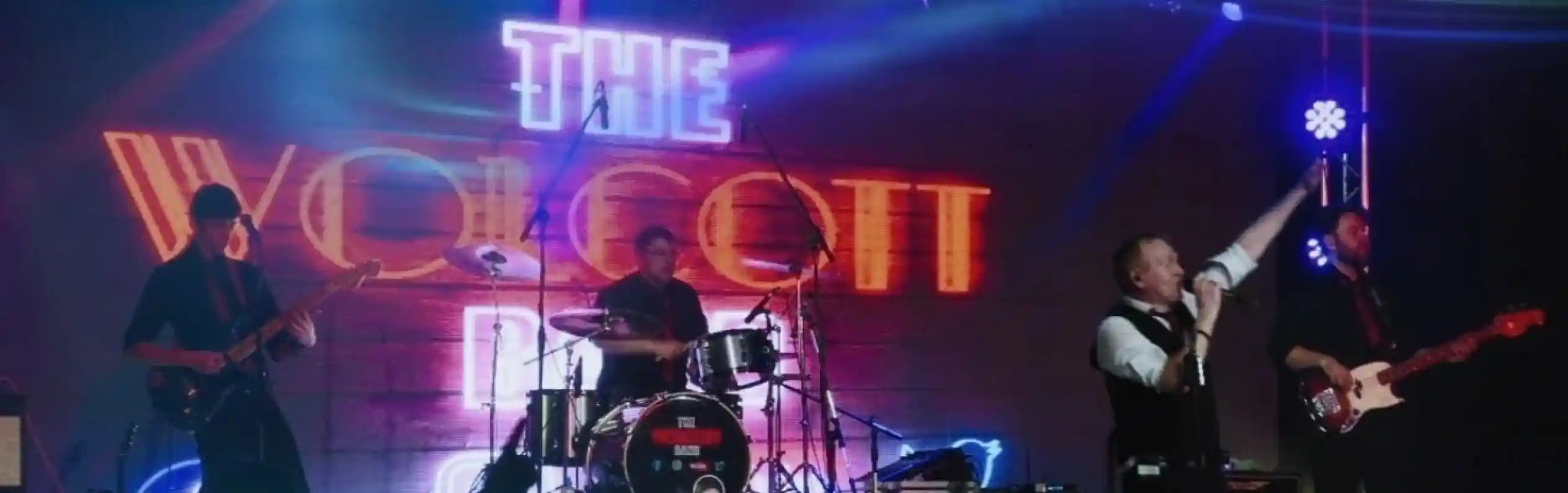 A live band performance with a lead singer on stage, energetically singing and gesturing. The band includes a drummer and two guitarists, all under colorful stage lights. The backdrop features the name "The Wolcott Band" in bright letters. Audience members are seen enjoying the performance.