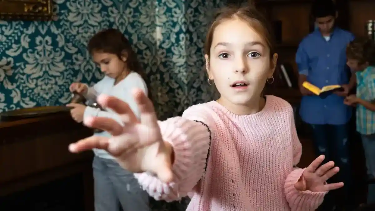 A group of four people stands in a colorful room filled with educational toys and decorations. A child holds a blue sign that says, "I'm still confused." The adults, smiling and looking at the child, convey a sense of playful engagement.