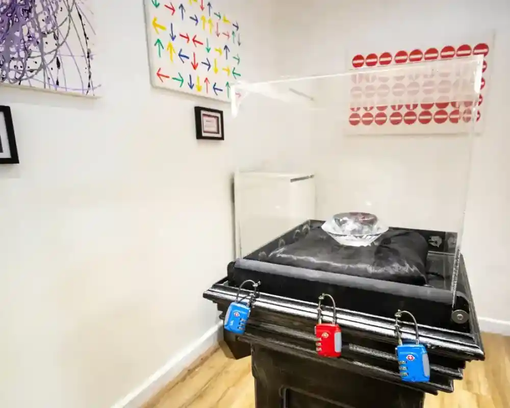 A clear display case surrounds a large, faceted crystal on a velvet cushion, positioned atop a black pedestal. Nearby walls feature colorful abstract art, including patterns of arrows and lip prints, framed in black. Three locks in blue and red secure the display.