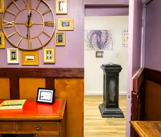 A purple-walled interior features a large wall clock and several framed pictures. A red-topped cabinet stands to the left, while a doorway on the right leads into a room with a pedestal displaying artwork. The floor is wooden.