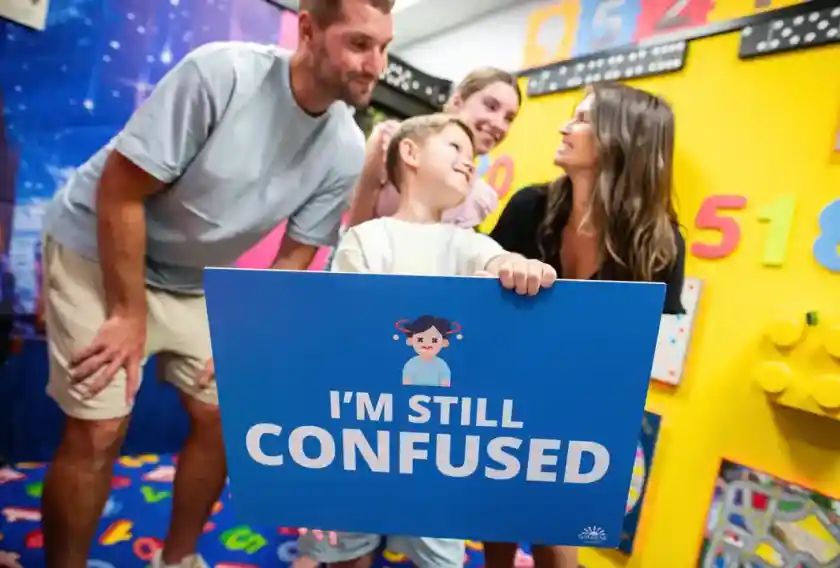 A group of four people stands in a colorful room filled with educational toys and decorations. A child holds a blue sign that says, "I'm still confused." The adults, smiling and looking at the child, convey a sense of playful engagement.