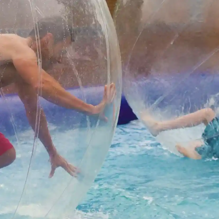 Two people are inside transparent inflatable balls, floating on water. One person, wearing red shorts, is upright and reaching out with one hand, while the other, in a blue swimsuit, is partially submerged and leaning back. Splashes of water surround them.