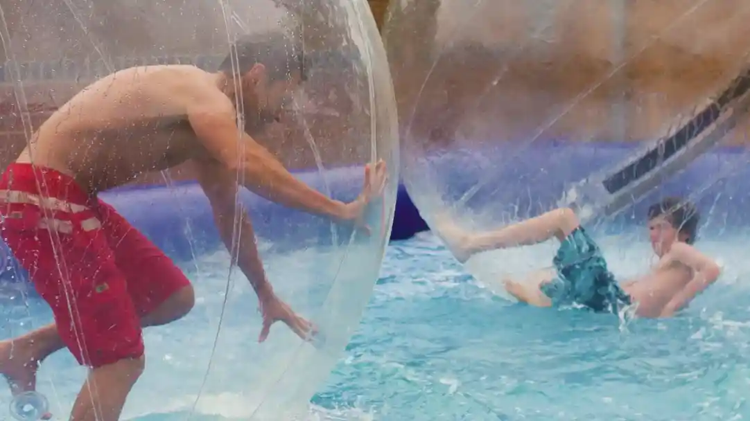 Two people are inside transparent inflatable balls, floating on water. One person, wearing red shorts, is upright and reaching out with one hand, while the other, in a blue swimsuit, is partially submerged and leaning back. Splashes of water surround them.