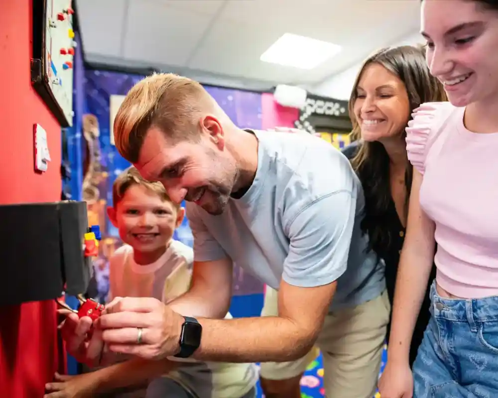 A man and woman assist a young boy with a colorful interactive activity on a red wall, while an older girl observes with a smile. The background features bright, playful decorations.