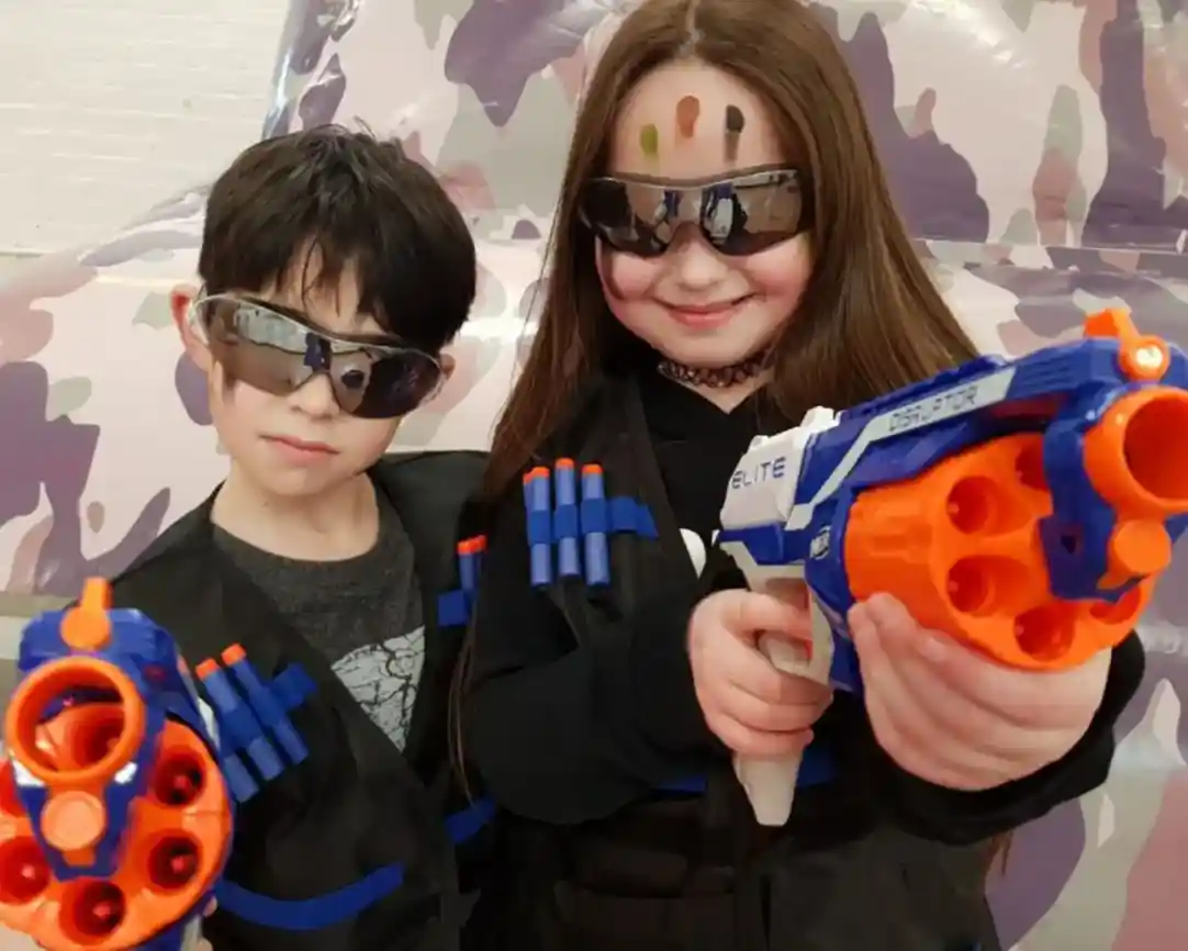 Two children dressed in tactical vests with toy guns. They wear sunglasses and have colourful face paint on their foreheads. The background features a camouflage design.
