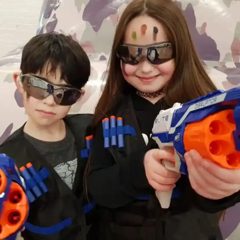 Two children dressed in tactical vests with toy guns. They wear sunglasses and have colourful face paint on their foreheads. The background features a camouflage design.