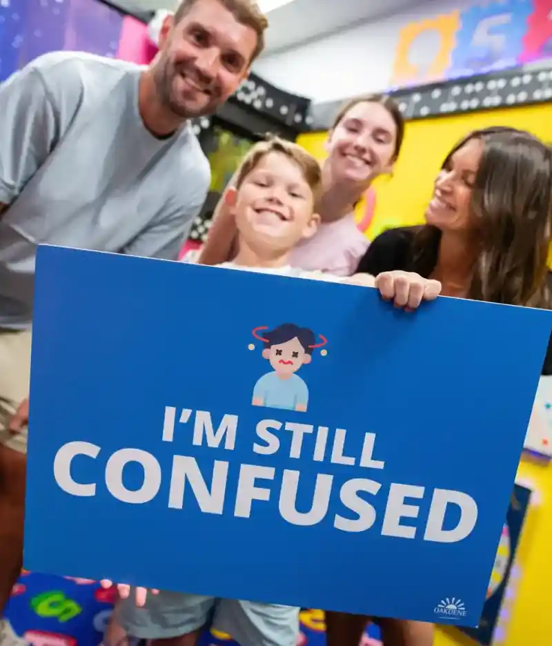 A couple stands on a balcony with a modern home in the background. The man and woman are smiling, dressed casually, with green grass and a landscaped area surrounding the house. The sky is clear and blue.