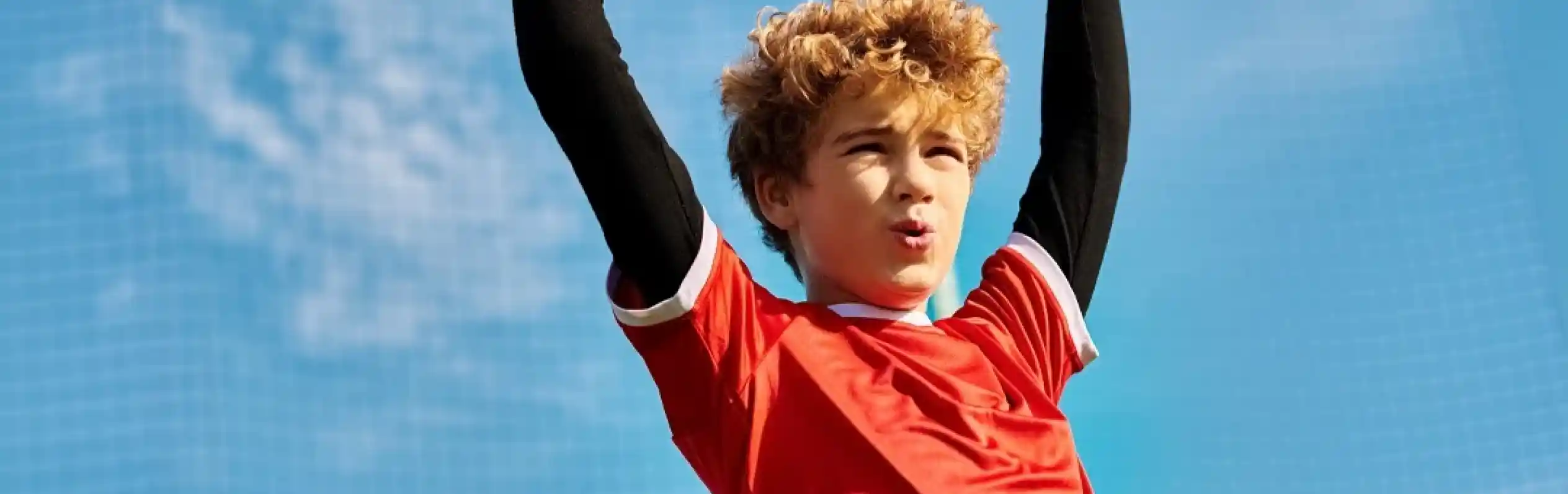 A young boy with curly hair raises his arms in celebration while wearing a red soccer jersey. He stands against a blue sky and soccer goal in the background.