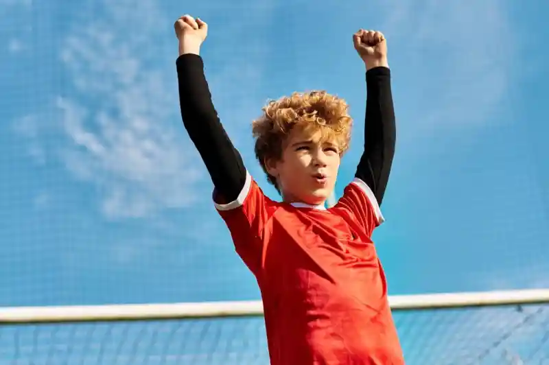 A young boy with curly hair raises his arms in celebration while wearing a red soccer jersey. He stands against a blue sky and soccer goal in the background.