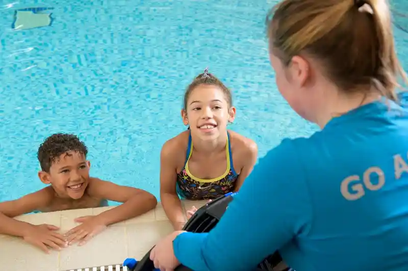 Two children in swimsuits smile and talk with a swimming instructor at the edge of an indoor pool. The instructor, wearing a blue shirt with 