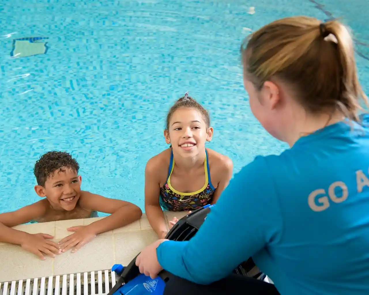 Two children in swimsuits smile and talk with a swimming instructor at the edge of an indoor pool. The instructor, wearing a blue shirt with 