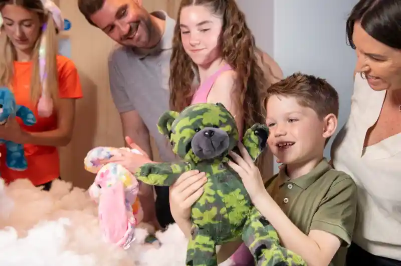 A smiling boy holds up a green camouflage teddy bear he has just made during a Stuff a Bear activity. Family members and staff watch happily, surrounded by colourful plush toys and stuffing.