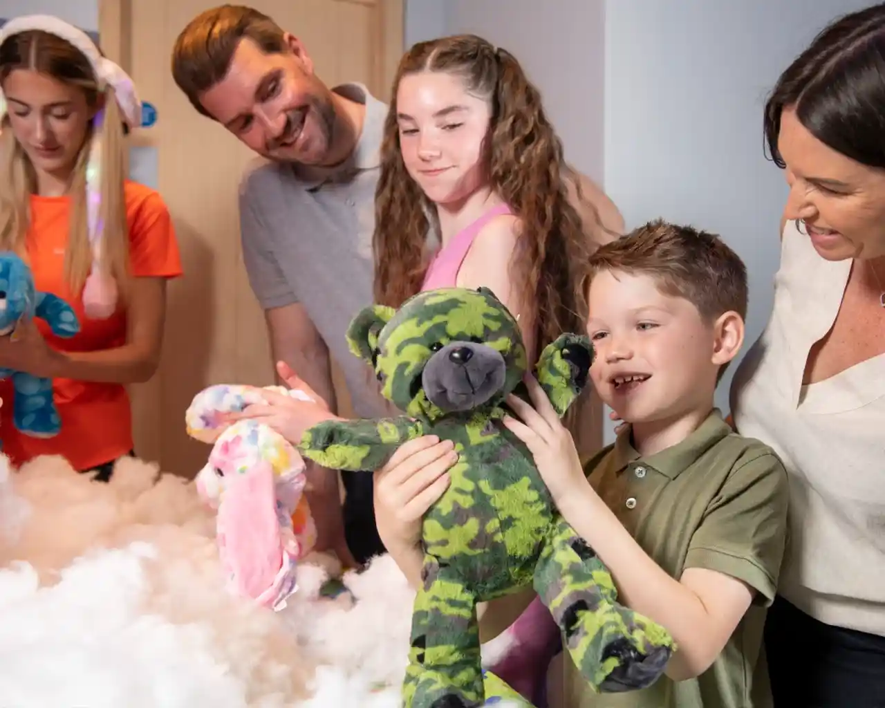 A smiling boy holds up a green camouflage teddy bear he has just made during a Stuff a Bear activity. Family members and staff watch happily, surrounded by colourful plush toys and stuffing.